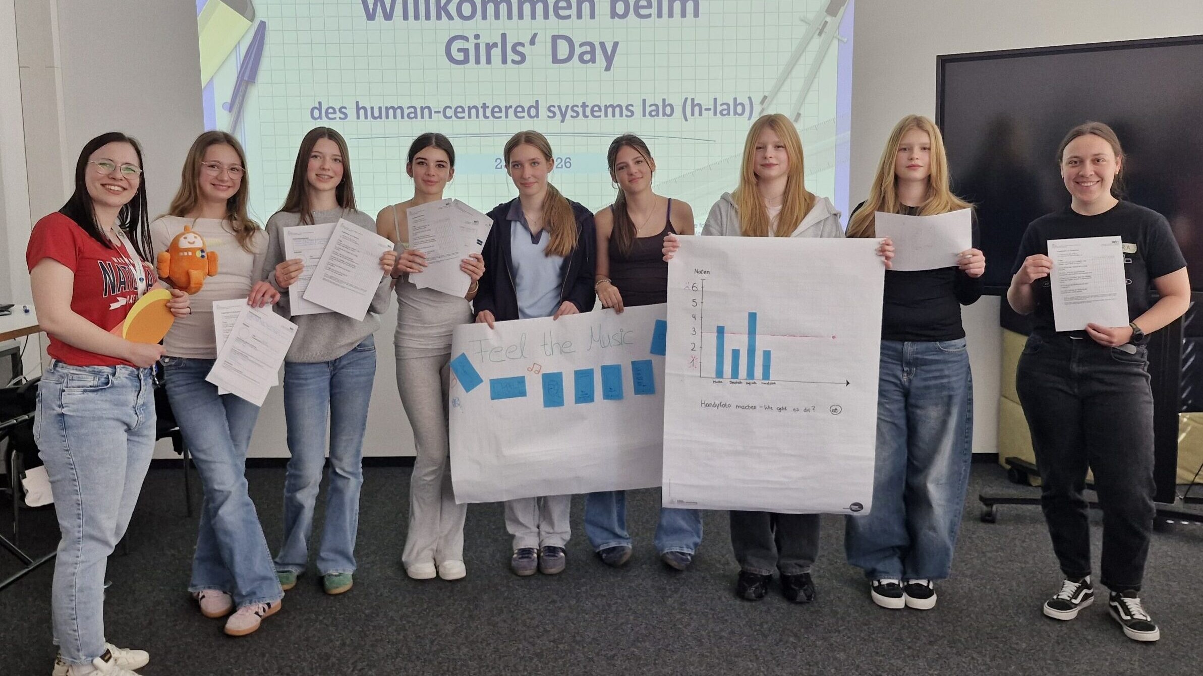 Gruppenbild mit neun Personen in einer Reihe. Ganz links Sandra Müller vom Human-Centered SysteDie Teilnehmerinnen des Girls' Day entwickelten eine App, die Emotionen selbstständig erkennt. Angeleitet wurden sie von Sandra Müller (ganz links ) und Chiara Krisam (ganz rechts) vom Human-Centered Systems Lab. © Human-Centered Systems Labms Lab, ganz rechts Chiara Krisam vom Human-Centered Systems Lab, dazwischen sieben der zwölf Teilnehmerinnen. Sie halten Poster mit einem Diagramm und einer Visualisierung der App hoch.