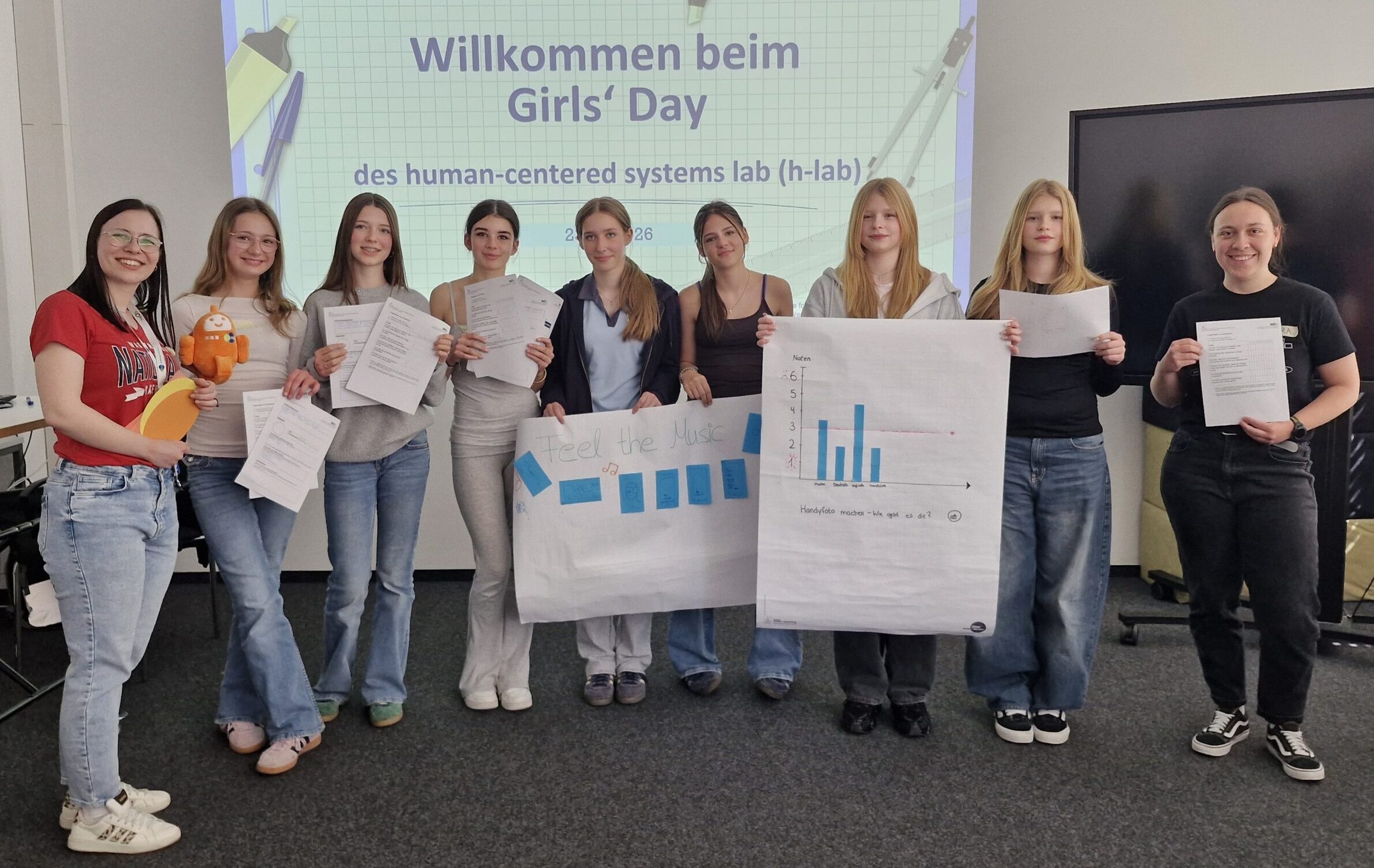 Gruppenbild mit neun Personen in einer Reihe. Ganz links Sandra Müller vom Human-Centered SysteDie Teilnehmerinnen des Girls' Day entwickelten eine App, die Emotionen selbstständig erkennt. Angeleitet wurden sie von Sandra Müller (ganz links ) und Chiara Krisam (ganz rechts) vom Human-Centered Systems Lab. © Human-Centered Systems Labms Lab, ganz rechts Chiara Krisam vom Human-Centered Systems Lab, dazwischen sieben der zwölf Teilnehmerinnen. Sie halten Poster mit einem Diagramm und einer Visualisierung der App hoch.