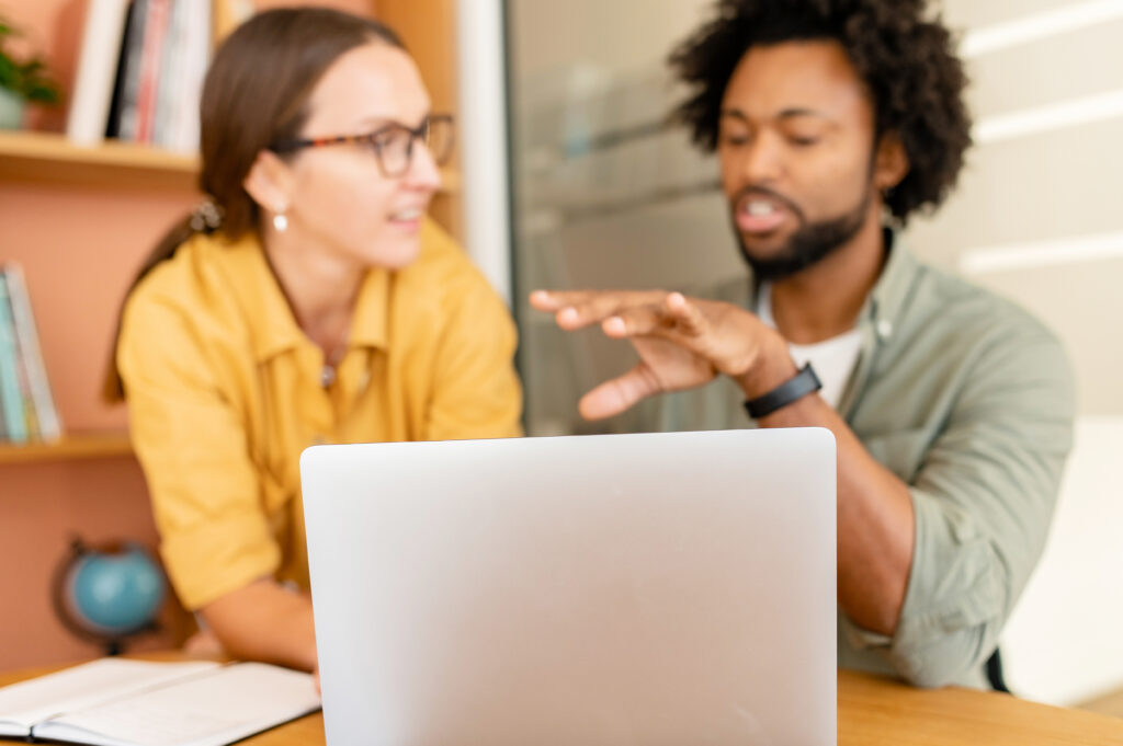 Scharf vorne ein Laptop von hinten, unscharf im Hintergrund zwei Personen im Gespräch, links eine Frau mit Brille und braunen Haaren, rechts ein Mann mit Locken und kurzem Vollbart, der mit seiner linken Hand gestikuliert.